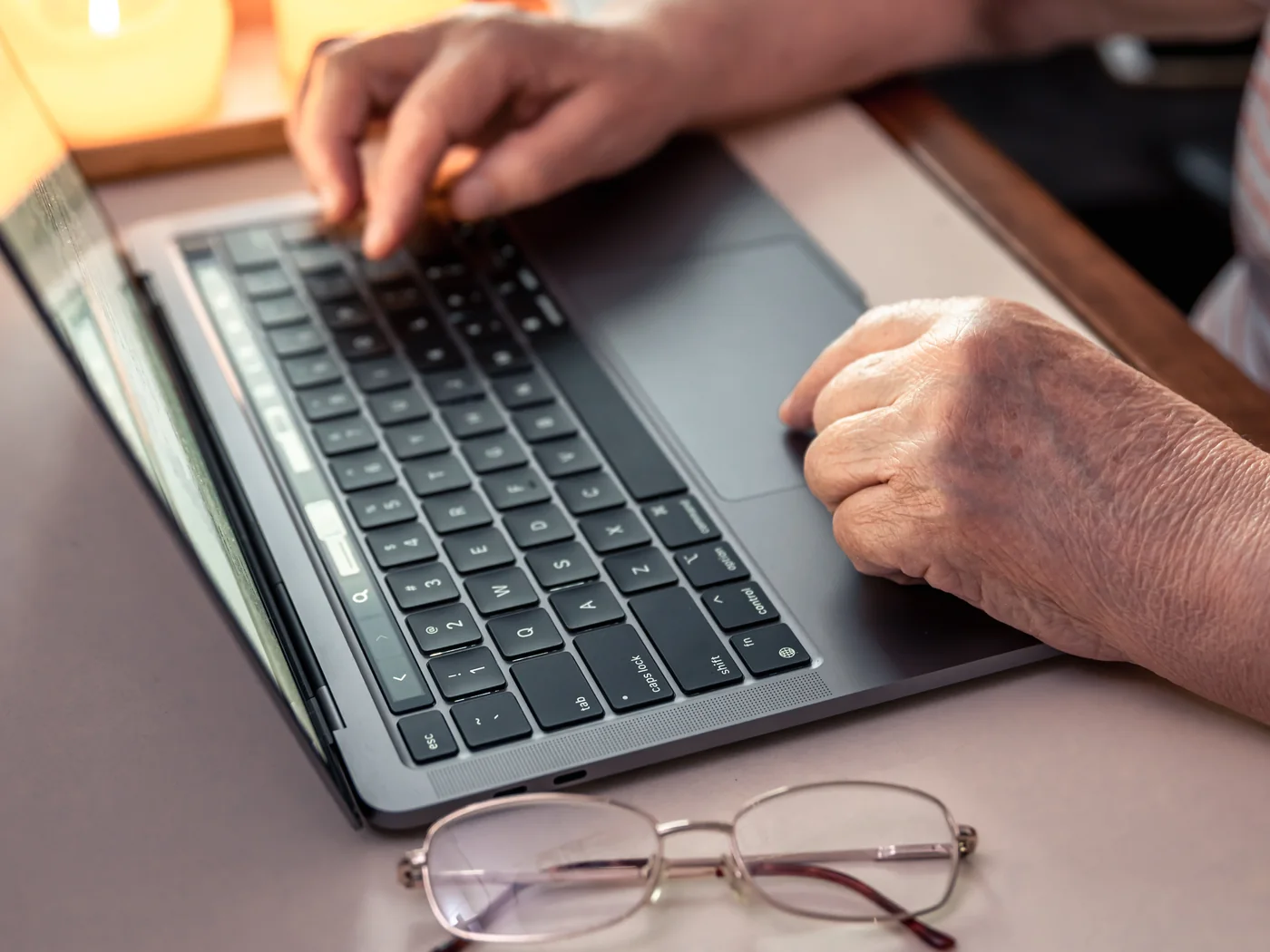 Hände einer älteren Person am eigenen Laptop, daneben die Lesebrille